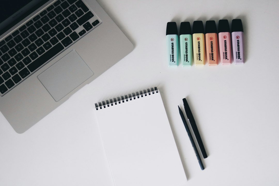 A laptop, a spiral notebook, two pens, and six colorful highlighters on a desk